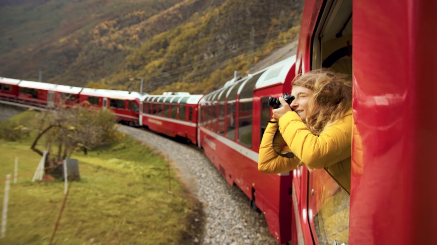 Inspiring female traveler and travel blogger look out of window of tourist train in cinematic beautiful mountains. Young beautiful woman hang out of window of red train. Excited world explorer Royalty-Free Stock Footage #1050828124