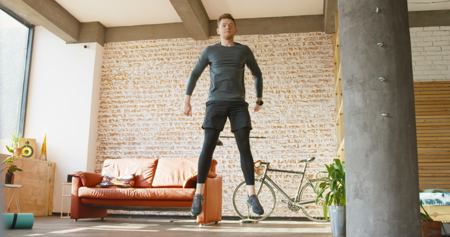 Caucasian young man with red hair in sporty style doing squats and jumping at home during quarantine. Self-isolated concept. Guy working out in apartment. Male keeping body in good shape.