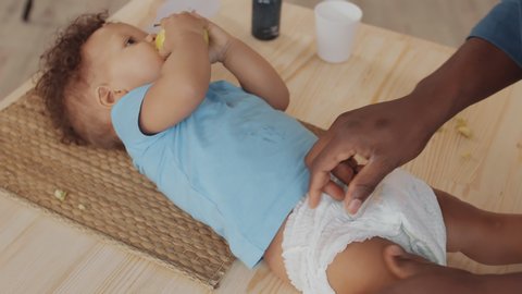 High angle view of male hands putting new disposable diaper on little African boy lying on wooden table and eating apple