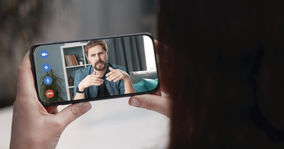 Back view of young girl sitting at home and having video chat with coworker on smartphone. Woman talking with man online during quarantine time. Focus on phone screen.