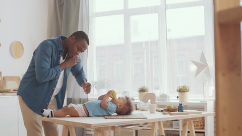 Medium shot of African man putting new fresh diaper on cute African toddler lying on wooden table with apple in his hands while talking on telephone