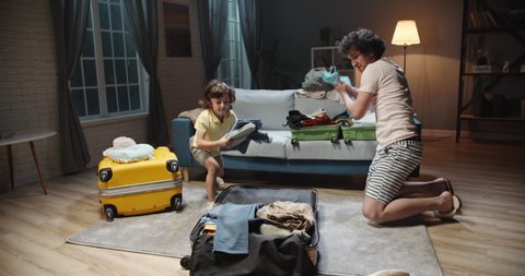 Funny asian young dad and preschooler son with curly hair are rapidly packing clothes in suitcase at home, preparing for their vacation. Family before road trip 4k footage