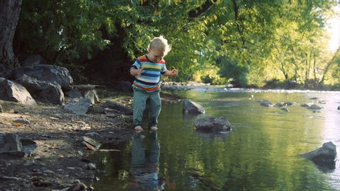 Toddler happily steps in river and starts to splash at sunset in slow motion