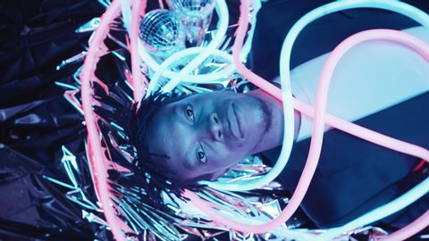 Top view studio shot of young handsome African American with braids man lying on metallic foil with glowing multicolored neon tubes and disco balls, looking at camera and smiling