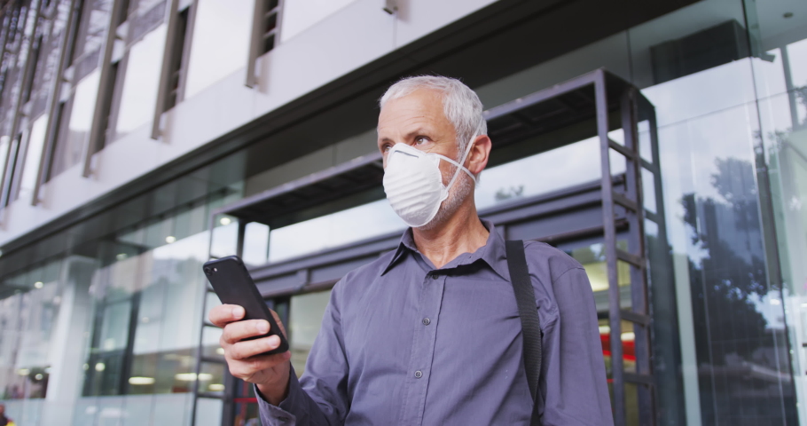 Middle aged Caucasian man out and about in the city streets during the day, wearing a face mask against coronavirus, covid 19 and using a smartphone, in slow motion.
