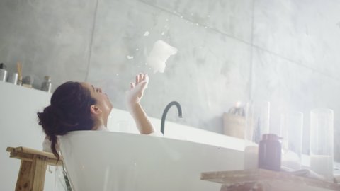 Close up cheerful woman blowing foam in light bathroo imn slow motion. Back view of smiling girl relaxing in bathtub at home. Relaxed woman having fun with foam in luxury bath.