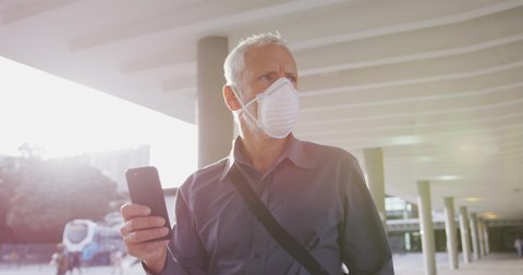 Middle aged Caucasian man out and about in the city streets during the day, wearing a face mask against coronavirus, covid 19 and using his smartphone, in slow motion.