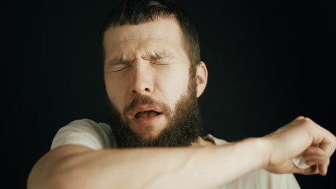 Handsome man with beard sneezes in the elbow. Bearded man is sneezing with his eyes closed on black background. Allergic sneezing or sneeze as sign of sickness. Close up portrait slow motion 4K video.