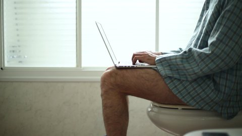 Relaxed man in a pijama working online with a laptop from the toilet at home 