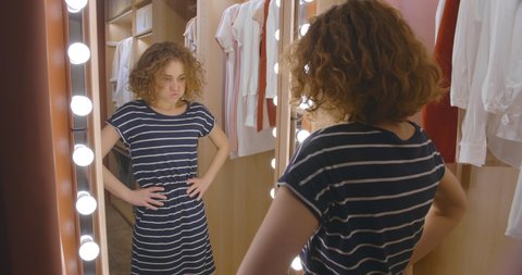 Young unhappy woman trying on dress to wear in front of mirror in wardrobe unsatisfied with her reflection. Distressed young girl dislike her appearance in mirror
