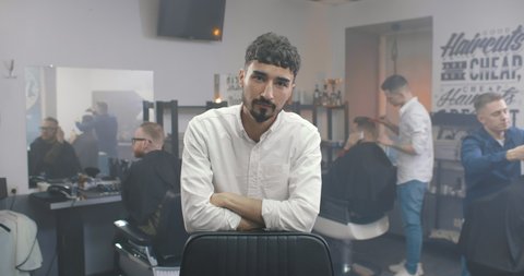 Portrait of bearded young barber in white shirt leaning in chair and looking at camera with colleagues working with clients in background in modern barber shop