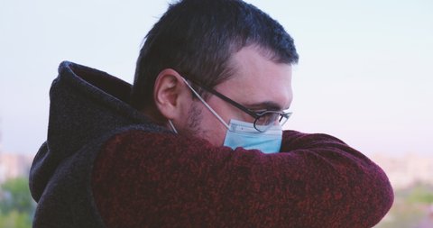 Man sneezes into his elbow, then takes a deep breath - side view. Coronavirus concept. Adult with glasses and short hair, wearing a medical mask and a hoodie.