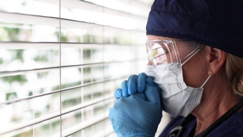 Stressed Doctor or Nurse On Break Praying At Window Wearing Face Mask and Protective Glasses.