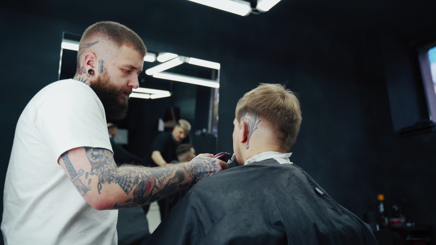 Barber trimming bearded man with shaving machine in barbershop. Hairstyling process. Close-up of a Hairstylist cutting the beard of a bearded male. | Shutterstock HD Video #1050908857