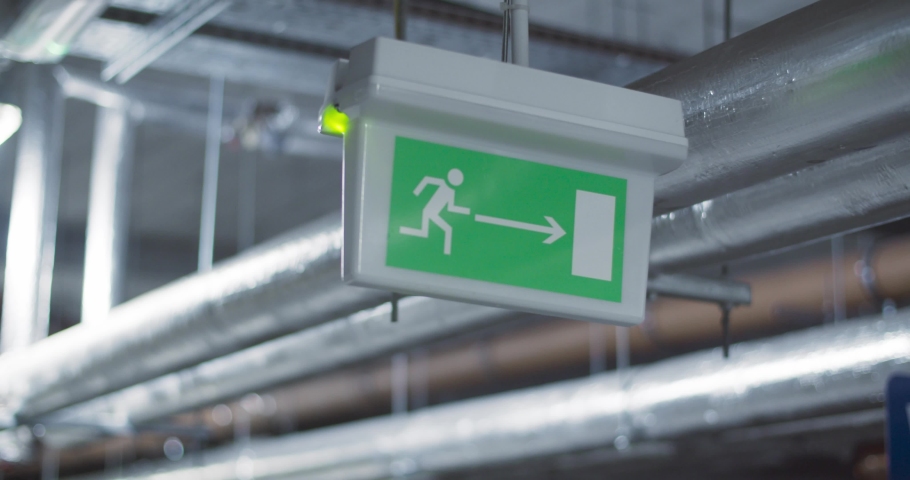 Green emergency, fire exit sign. Standard international symbol safe exit sign is hanging from the ceiling at underground garage. Industrial pipes on the background Royalty-Free Stock Footage #1050912811