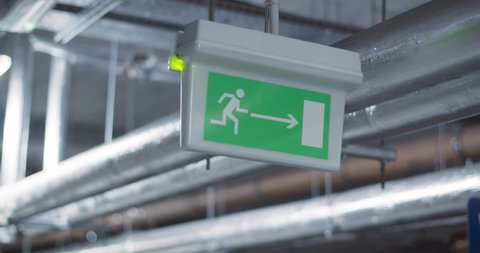 Green emergency, fire exit sign. Standard international symbol safe exit sign is hanging from the ceiling at underground garage. Industrial pipes on the background
