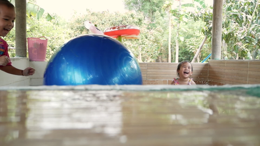 Cute little Asian child girl playing a blue gym ball with her sister in swimming pool at backyard. Happy lifestyle with family on vacation day.