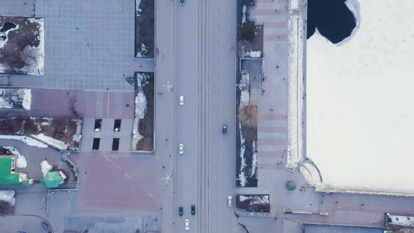 Yekaterinburg, Russia. The historic Plotinka dam on the Iset River, separating the central city pond from the lower waterway, viewed from directly above in a vertical aerial perspective. The dam structure spans the full width of the frame, with a paved pedestrian promenade running along its length and a multi-lane road carrying traffic across the water. To the right, the frozen central pond extends toward the edge of the frame, its ice surface broken by a dark, irregular patch of open water. To the left, the lower river channel flows through a series of rectangular basins and sluice structures that form the spillway system, with water visible cascading through the concrete channels. The surrounding area features geometric stone-paved squares, patches of melting snow, and sparse winter vegetation. The granite-faced dam has served continuously since the 1720s, making it one of the oldest unreinforced hydraulic structures of its kind in continuous operation. The cool, diffuse daylight and muted blue-grey palette emphasize the late winter or early spring season. HEAD OVER SHOT