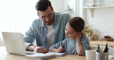 Young father checking homework helping cute school child daughter with studies sit at kitchen table. Adult parent or tutor explaining kid girl distance learning at home. Private education concept