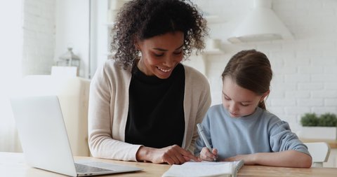 Smiling african woman tutor or foster parent mum helping cute caucasian school child girl doing homework sitting at kitchen table. Diverse nanny and kid learning writing in notebook studying at home.