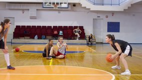 Two young female athletes in sportswear practicing dribbling and passing a basketball on court while their teammates watching their workout and recording video with smartphone