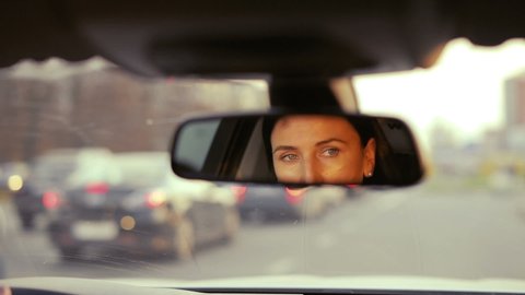 Close up of a reflection of a woman in a rearview mirror of a car while driving in a traffic jam in the evening. Beautiful woman driving a car.
