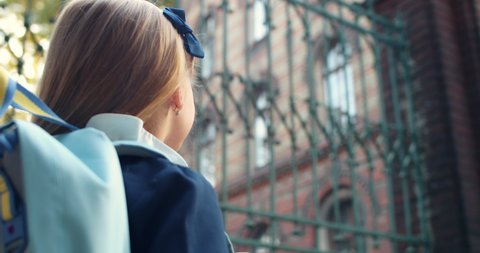 Backside view of little schoolgirl standing and looking at school. Child with backpack afraid of going to school. Concept of bullying among adolescents and schoolchildren