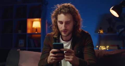 Young hipster guy making yes gesture and smiling while having good news. Handsome long haired man winning while using his smartphone and sitting on couch at home. Concept of success