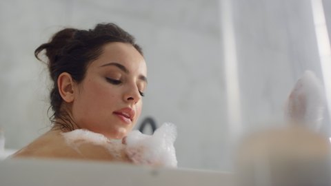 Close up sensual woman blowing foam in bathtub slow motion. Bottom view of romantic girl touching hands with foam in bath at home. Sexy woman resting in bath with bubbles indoors.