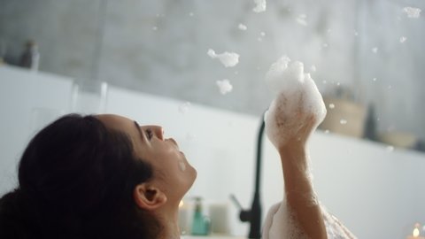 Close up relaxed woman blowing foam in luxury bathroom in slow motion. Back view of happy girl having fun with foam in modern bath. Sensual woman relaxing in bathtub at home.