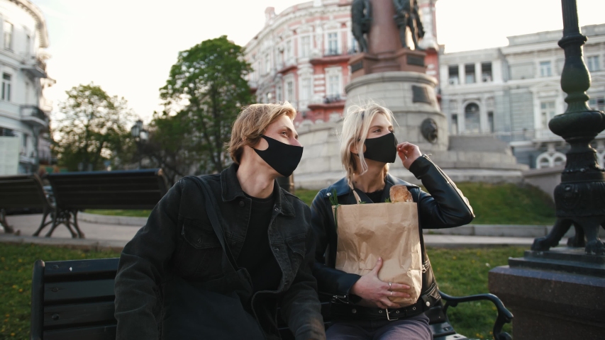 Portrait of happy young couple in protective masks sitting on a bench outdoors in old city center and talking to each other, slow motion