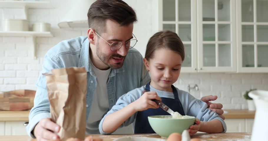 Caring dad teaching child daughter learning making dough in kitchen. Young adult daddy embracing cute little kid girl wearing apron helping parent father preparing cake, pie or pancakes at home. Royalty-Free Stock Footage #1050958684