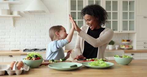 Cheerful mixed race young mom and child daughter giving high five cooking together in kitchen. Happy african foster parent mother, nanny, teaching cute kid having fun making healthy salad at home.