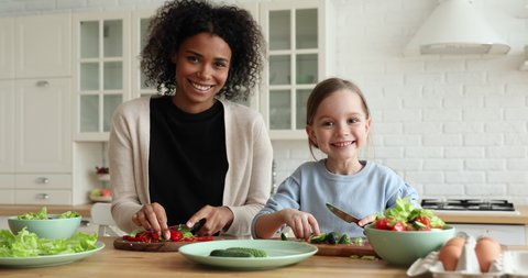 Happy mixed race vegan family young foster african mom and cute small caucasian adopted child daughter enjoy cooking together, preparing healthy organic vegetable salad, looking at camera. Portrait.