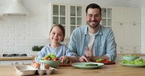 Happy cute small kid daughter cooking with father looking at camera stand at kitchen table. Young dad teaching child learning preparing healthy food cutting vegetable salad. Vegan family making meal.