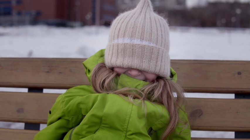 A little happy girl pulls a knitted beige hat over her eyes. A faceless child laughs hiding behind a cap,kid sitting on a wooden swing against a background of white snow. | Shutterstock HD Video #1050960064