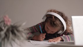 Small blonde kid doing homework by video call in front of a laptop using headphones, a pencil and a rubber at home