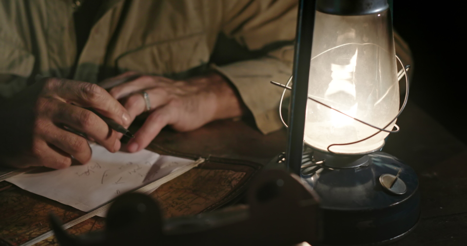 Military man writing strategy of defeating enemy. Tilt up of World War II soldier sitting at table with burning kerosene lamp and taking notes of strategy while sitting in enemy rear Royalty-Free Stock Footage #1050968596