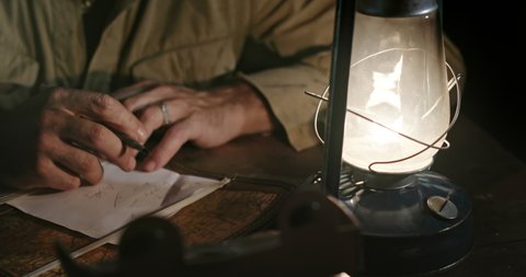 Military man writing strategy of defeating enemy. Tilt up of World War II soldier sitting at table with burning kerosene lamp and taking notes of strategy while sitting in enemy rear