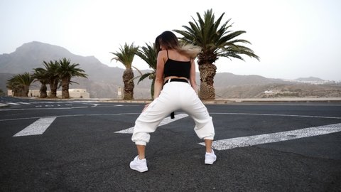 Hot girl in white is dancing outdoor at the road with tropical trees and hills on background. Sexy long haired brunette woman is practicing dance, moving her body professionally