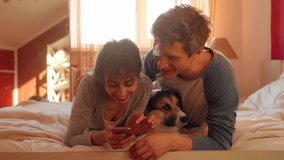 lovely couple lying on bed in bedroom with cute Welsh Corgi dog, holding smartphone, watching photo or video online together. Concept stay at home, friendship with pet, spending time together.