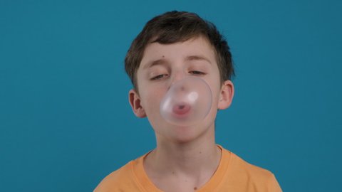 Happy boy chewing gum and learning to inflate bubbles and bursting it joyfully smiling looking at camera slow motion on blue background. Child. Childhood. Emotions of people. Life style