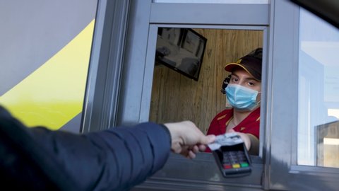 MINSK, BELARUS - April 8, 2020: Visitor orders food and pays by credit card at McDonald's drive thru during pandemic. Cashier in a medical mask for protection against viruses.