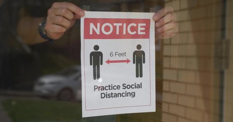 A man puts a social distance sign on the front door of his shop. Social or physical distancing was a recommended procedure to help slow the spread of COVID-19 during the pandemic of 2020.	