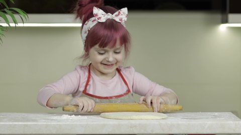 Cooking homemade pizza. Little child girl in the kitchen dressed in apron and scarf like chef. Kid roll dough with rolling pin, smiles happy. Concept of: nutrition, cooking school, education, game
