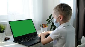 Cute little school boy in medical mask learning on computer, distance education concept. View from back, Green screen