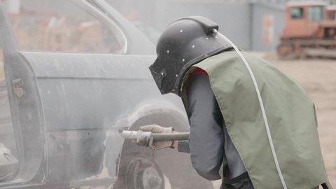 Worker sandblasting metal surface of car. Sandblasting of metal surface of the car by compressed air of the sandblaster gun