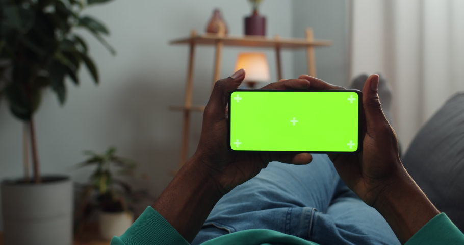 Man holding green screen smartphone in horizontal landscape mode with trecking markers while lyying on sofa . African guy watching video while looking at mock up screen Royalty-Free Stock Footage #1051054909