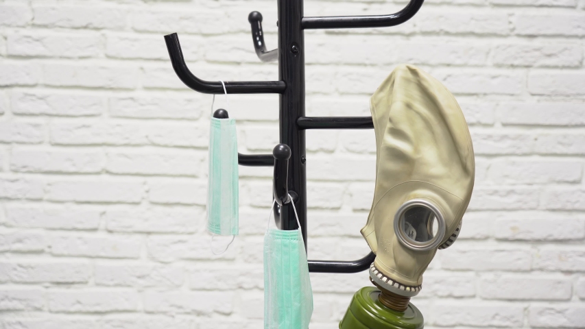 Hands of man takes an old protective military gas mask on background of white brick wall. Guy puts on rubber helmet on black hanger with two medical masks. Covid-2019 virus protection concept.