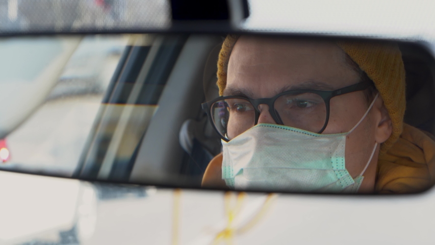 Handsome Caucasian man driver in a turquoise protective medical mask with black glasses and a yellow hat looks in the rearview mirror and ride a car close up. Any cars passing by. Inside view.
